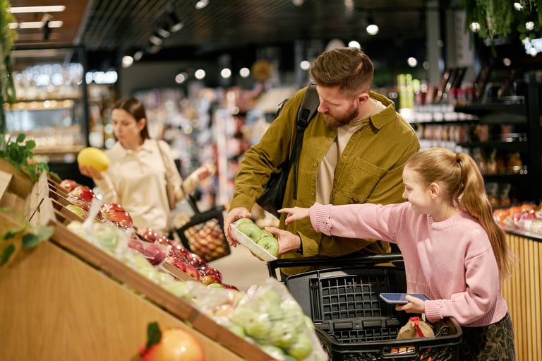 Family exploring produce section in grocery store while selecting fresh vegetables. Shelves stocked with colorful fruits and vegetables visible in background creating lively scene